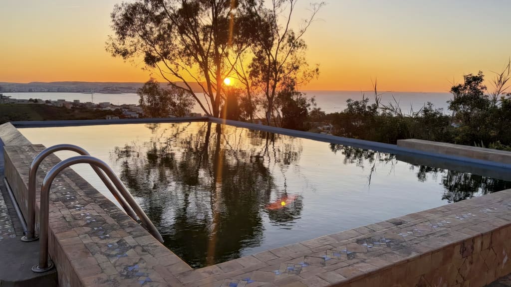 Piscine à débordement au coucher du soleil, margelle en zellige, avec la baie de Tanger et la Méditerranée à l'horizon.
