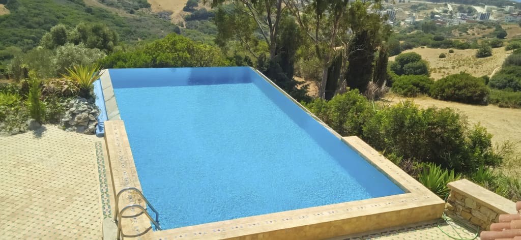 Piscine à débordement d'un bleu vif bordée de zellige, entourée de végétation et dominant les collines.